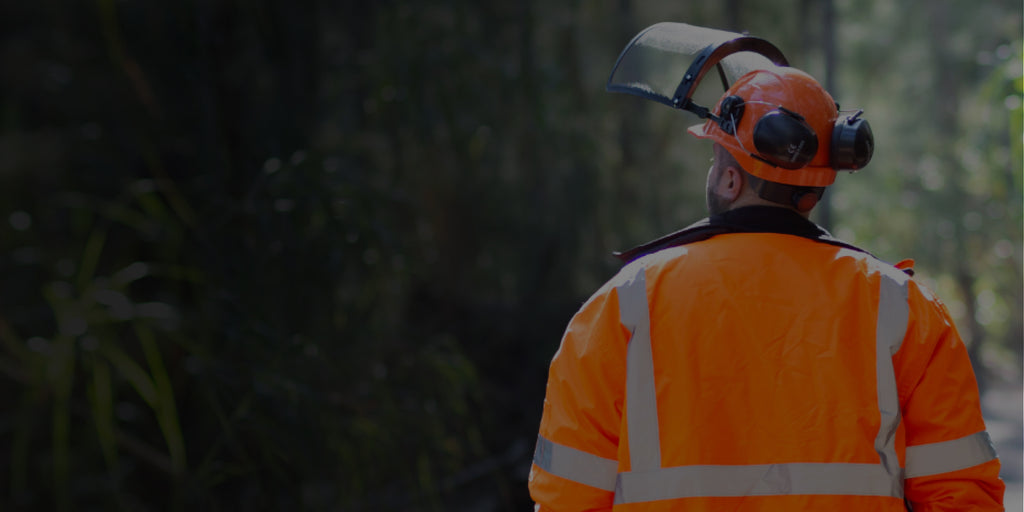 Male arborist wearing protective gear, including an orange hard hat with black face shield and earmuffs, as well as an orange high visibility safety jacket. He is walking through a path of trees