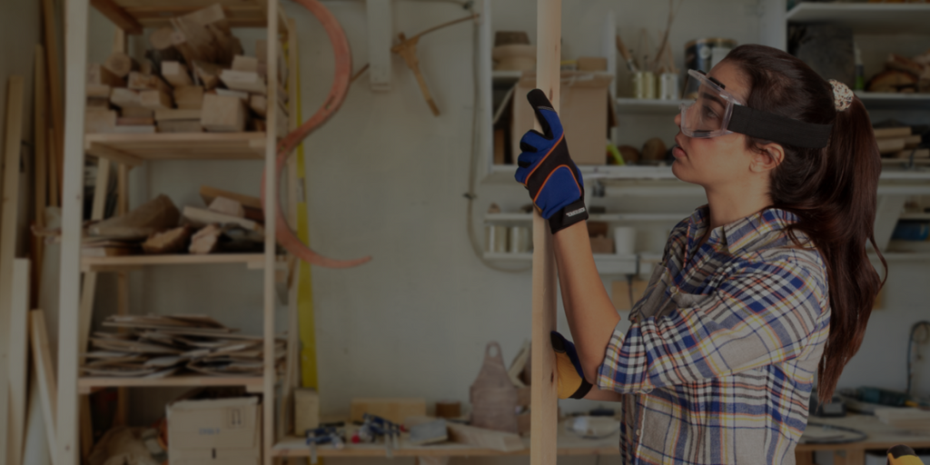 young woman in in her DIY woodworking workshop, she is wearing blue jorestech touchscreen safety work gloves and ventilated goggles while examining a plank of wood