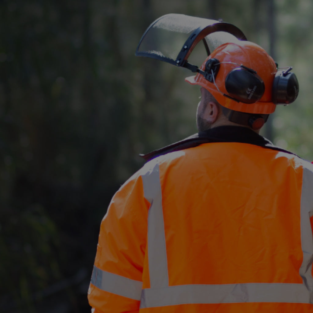 Male arborist wearing protective gear, including an orange hard hat with black face shield and earmuffs, as well as an orange high visibility safety jacket. He is walking through a path of trees