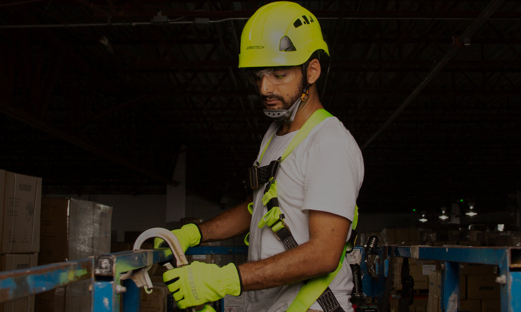 Young man working at high elevations equipped with safety gear to keep him safe from falls. He is wearing a work-at-height hard hat with ventilated side slots, headlamp mounting clips, and universal earmuff mounting slots. He is also wearing a full body safety harness and work gloves made of high visibility yellow fabric. He is being kept safe thanks to a fall arrest system comprised of a lanyard that would stop a fall and keep him safe if he were to slip from high altitudes. 