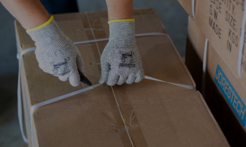 Grey Jorestech cut-resistant HDPE gloves with coated palms, used for worker safety in a warehouse work setting. The safety gloves are being used to protect the hand from cuts while he cuts a box open with a sharp knife. 