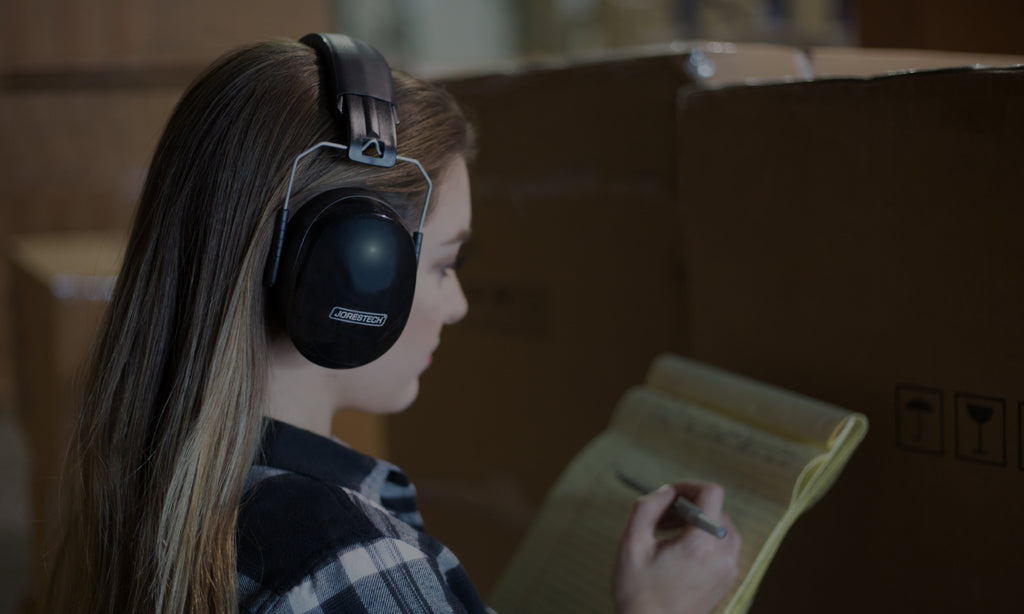 Warehouse worker doing inventory near loud equipment. She is protecting her ears with a set of Jorestech earmuffs, which provide 27dB (decibels) of sound suppression and a noise reduction rating of SNR 31dB (decibels).