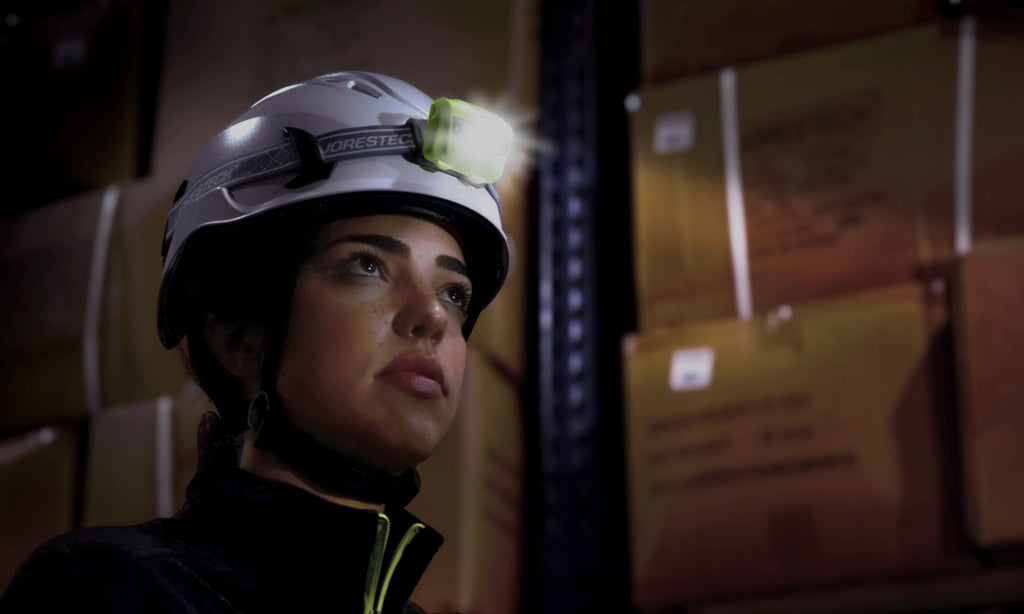 Warehouse worker wearing the jorestech climbing hard hat with ventilated slots. This work-at-height helmet has mounting clips that are used to mount headlamps and other accessories directly onto the safety hat. The wearer has a mounted headlamp, which she is using while she works in a low-light environment.