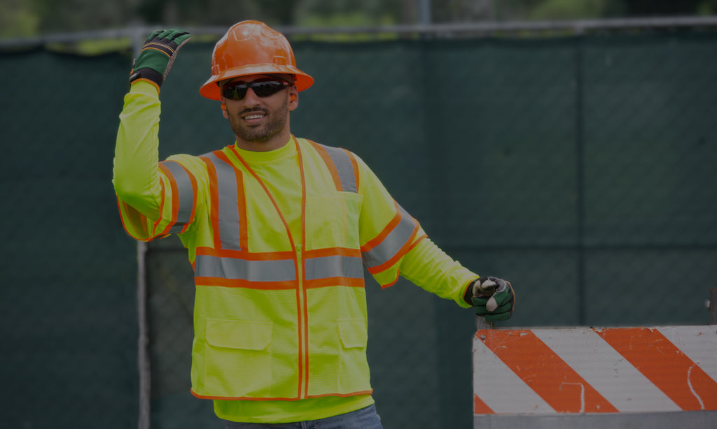 Roadside worker sporting a high-visibility safety look including an orange hard hat for head protection, green work gloves for hand protection, a high visibility yellow shirt, and a two-toned orange and yellow high visibility safety vest with ANSI compliant reflective stripes