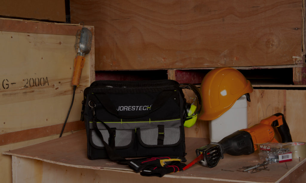 Jorestech organizer tool bag with a carpenter or builder's belongings scattered around the workplace. There is a cap-style hard hat and a saw, as well as safety earmuffs and work gloves to protect the worker's hands and ears.