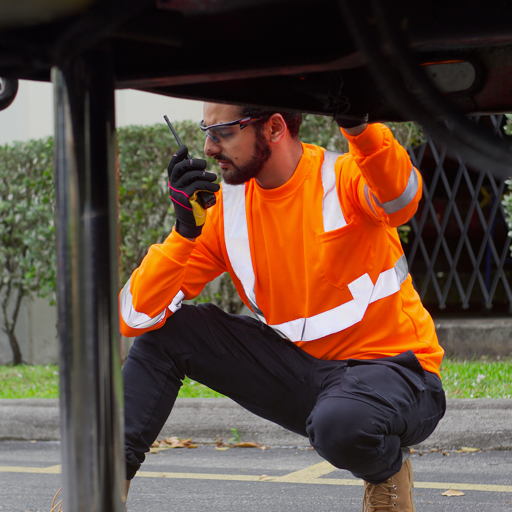 A worker wearing an orange reflective safety shirt talking on the portable radio beside a large truck