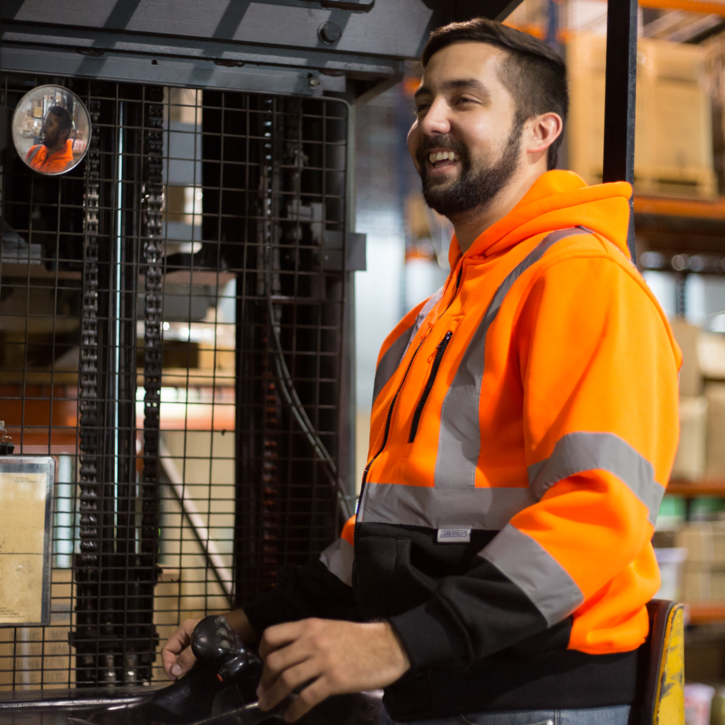 Image of a man wearing an orange JORESTECH safety sweatshirt with reflective stripes while he is driving a forklift inside a warehouse