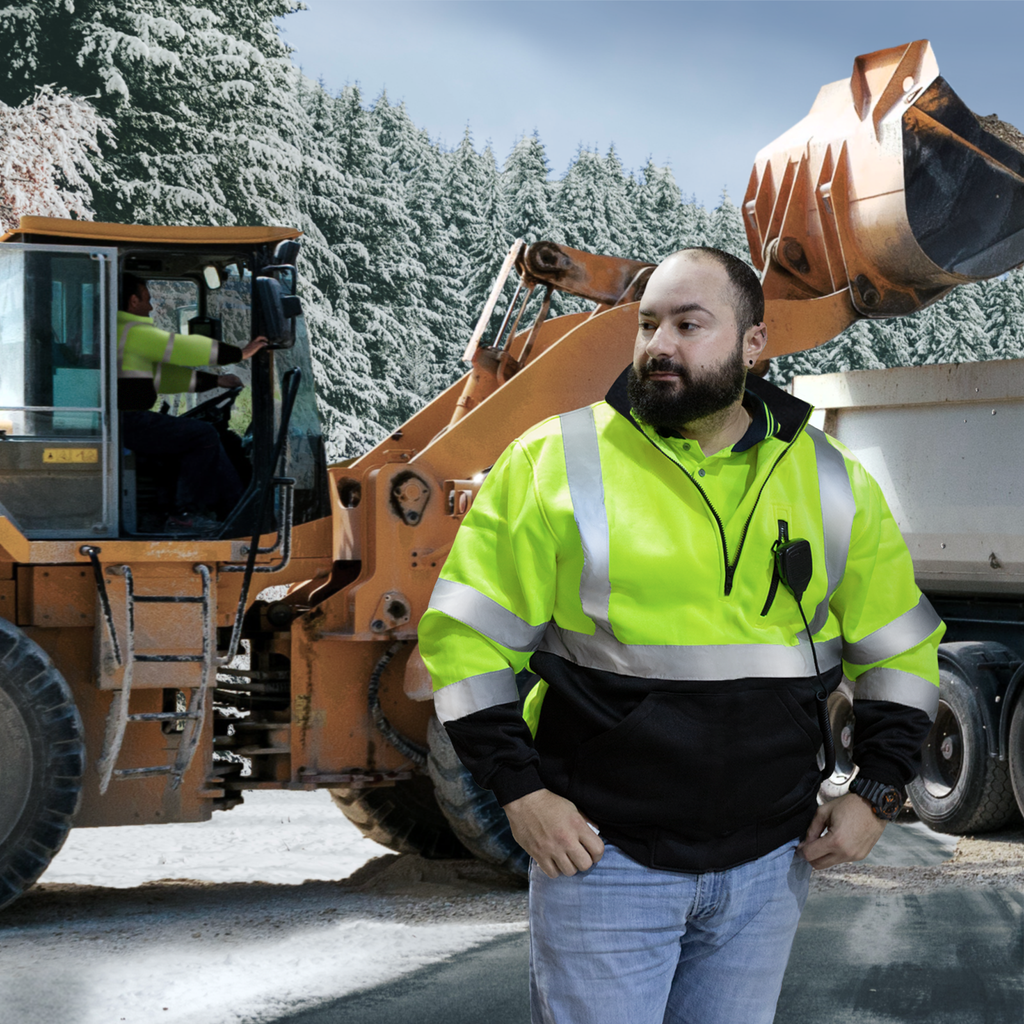 Image of man wearing the vi-vis yellow JORESTECH safety sweater with reflective stripes. The man is in a road construction during winter time and there are 2 large tucks behind him. The driver of one on the tucks is also wearing a yellow safety sweater